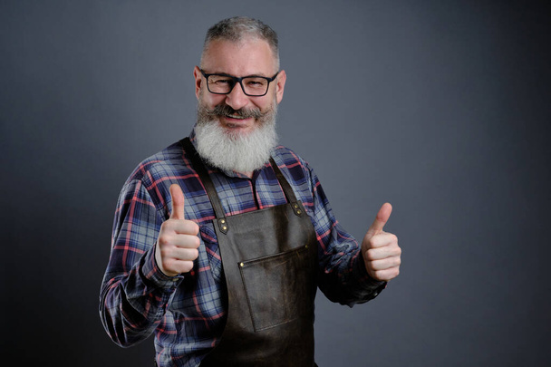 Портрет красивого взрослого бородатого мужчины, одетого в кожаный фартук, показывающего большие пальцы на сером фоне, белого рабочего с улыбкой на бороде - Фото, изображение