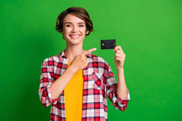 Photo of optimistic cute short hairstyle girl pointing card wear orange checkered shirt isolated over vivid green color background - Foto, Imagen