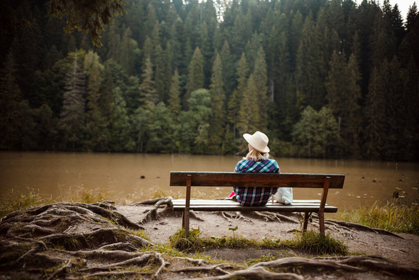Vista trasera de una joven con camisa y sombrero de mezclilla, sentada en un banco junto a un árbol con muchas raíces, admira el paisaje del lago, el bosque de pinos y el relieve de las montañas en el fondo, en el Lago Rojo, Cárpatos del Este, Rumania - Foto, Imagen