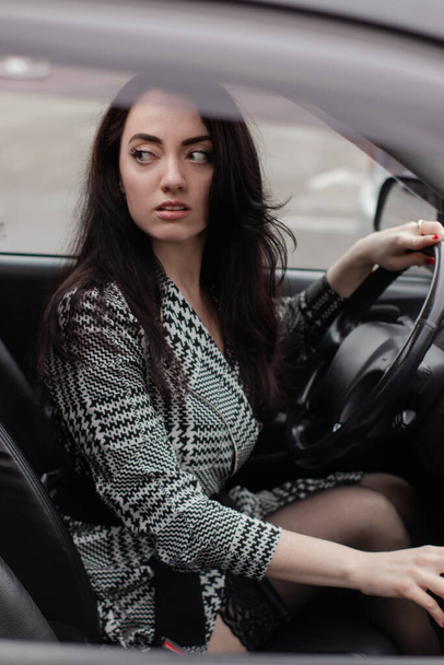 黒い車の中でグレーのチェッカーのドレスで魅力的なブルネットの肖像画。車の中の女の子。ビジネスウーマン - 写真・画像