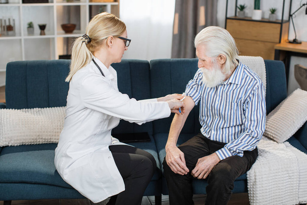 Mature professional confident blond woman doctor doing injection of vaccine or medicine to senior bearded male patient, while making visit him at home - Photo, image