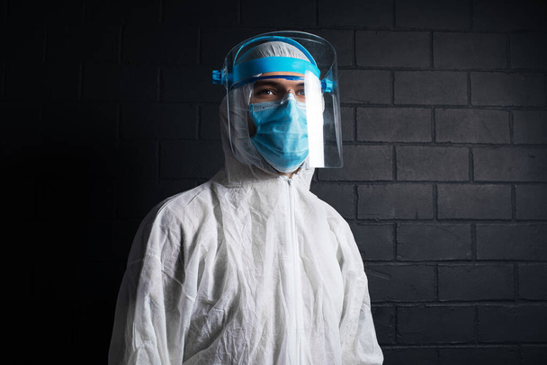 Studio donker portret van jonge arts in PPE-pak tegen coronavirus en covid-19. Achtergrond van zwarte bakstenen muur. - Foto, afbeelding