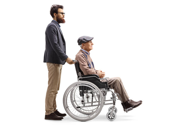 Full length profile shot of a bearded man pushing his elderly father seated in a wheelchair isolated on white background - Photo, image