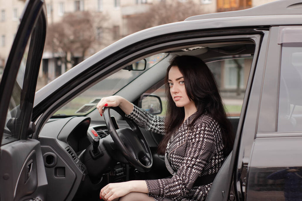 πορτρέτο μιας ελκυστικής μελαχρινής σε γκρι καρό φόρεμα σε μαύρο αυτοκίνητο. Κορίτσι με αυτοκίνητο. Επιχειρηματίας - Φωτογραφία, εικόνα