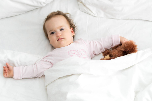 Auténtico retrato lindo bebé caucásico regordete niña o niño en rosa somnoliento al despertar con oso de peluche mirando a la cámara en la cama blanca. Cuidado de niños, Infancia, paternidad, concepto de estilo de vida - Foto, Imagen
