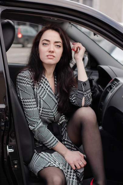 portrét atraktivní brunetky v šedých kostkovaných šatech v černém autě. dívka v autě. obchodní žena - Fotografie, Obrázek
