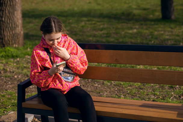 Une fille s'assoit sur un banc de parc et utilise un smartphone pour communiquer avec ses proches dans un chat vidéo. L'enfant écoute de la musique ou la voix des gens à l'aide d'écouteurs sans fil. Espace libre pour le texte - Photo, image