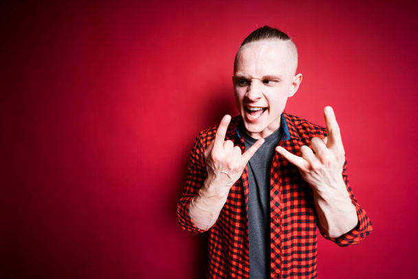 A man shows the Hookem horns gesture and shouts loudly against a red background in the studio. Rock music concept - Photo, Image