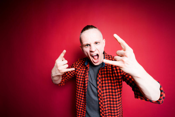A man shows the Hookem horns gesture and shouts loudly against a red background in the studio. Rock music concept - Photo, Image