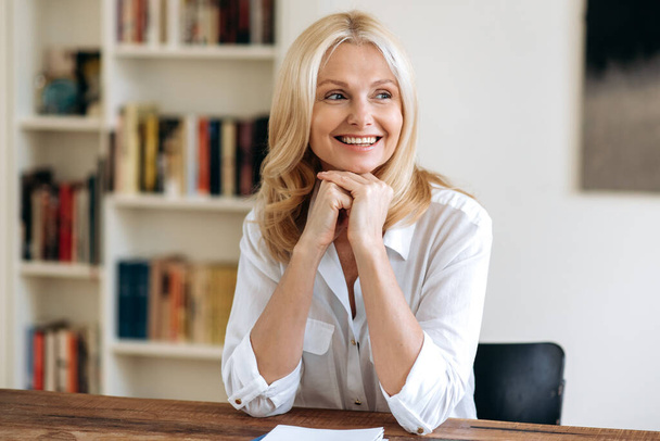 Portret van gelukkige elegante succesvolle volwassen blonde vrouw in wit stijlvol shirt, zit aan het bureau op kantoor, het nemen van een pauze van het werk, kijken naar de zijkant, denken, glimlachen vriendelijk - Foto, afbeelding