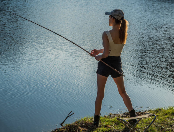 Uma menina fica na margem de um lago da floresta e captura peixes com uma vara de pesca. - Foto, Imagem