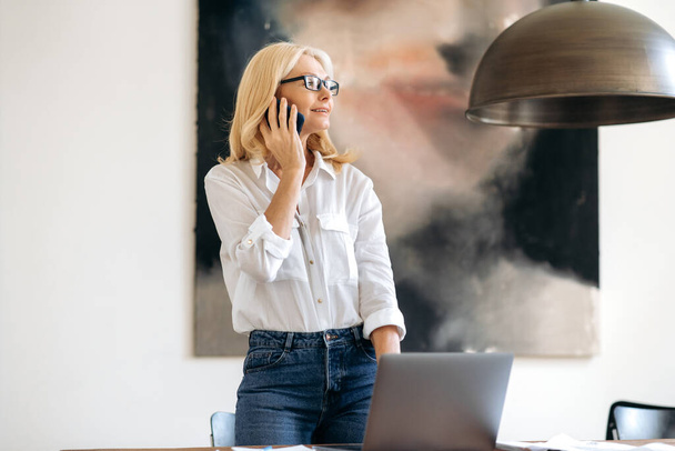 Beautiful successful confident senior blonde woman, stylishly dressed, business leader, broker or lawyer, standing near the desk, talking to employee or client on smartphone, smiling, looks away - Foto, immagini