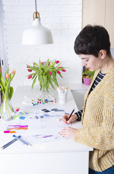 若いブルネットの女性は色鉛筆で描くファッションドレスを設計します。様々なカラフルなファッション図面やスケッチ - 写真・画像