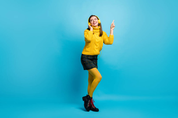 Full length photo of cute young lady wear yellow sweater headphones dancing pointing empty space isolated blue color background - Фото, изображение
