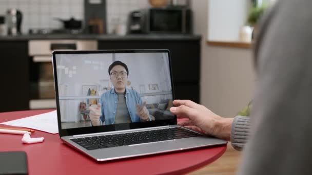 Přes rameno nepoznatelné osoby sedí u stolu s přenosným počítačem s spuštěním videokonference na mladého Asiata mluví - Záběry, video