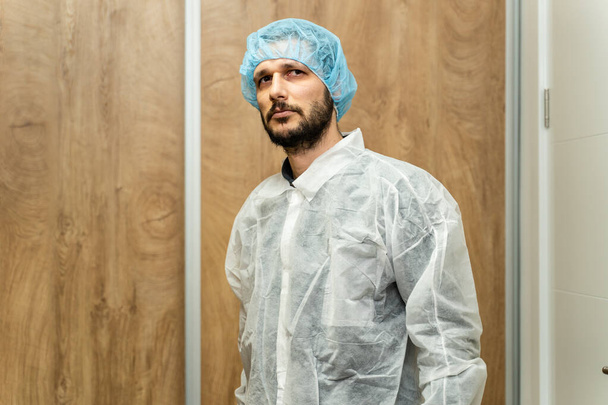 Πορτρέτο του καυκάσιου άνδρα που φοράει προστατευτική μάσκα και καπελάκι όχλου - Νεαρός άνδρας γιατρός ή επιστήμονας ετοιμάζεται για εργασία στο εργαστηριακό νοσοκομείο μπροστινή όψη με θέα στο χώρο αντιγραφής κάμερας - Φωτογραφία, εικόνα