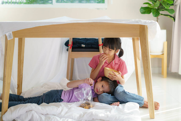 Asiático lindo niño alimentación snack a su hermana hermano mientras está sentado en un fuerte manta en sala de estar en casa para escondite perfecto lejos de sus otros miembros de la familia y para que jueguen imaginativamente. - Foto, imagen