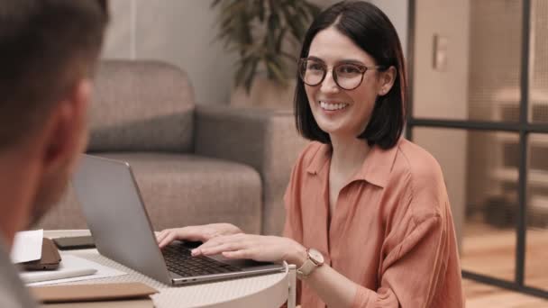 Waist-up of young Caucasian woman wearing glasses, typing on portable computer on coffee table, talking to unrecognizable person, smiling - Footage, Video