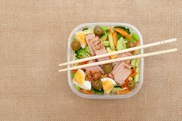 Healthy chicken salad with vegetables in plastic container with chopstick. Isolated on jute background. Top view. Copy space. - Photo, Image