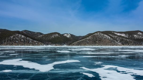 Na hladkém ledu zamrzlého jezera jsou vidět skvrny sněhu a prasklin. Na břehu, na pozadí modré oblohy, je zalesněné pohoří pokryté sněhem. Bajkal - Fotografie, Obrázek