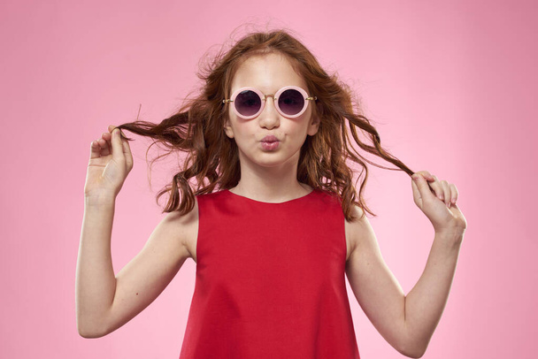 Schattig klein meisje rood jurk zonnebril studio roze achtergrond - Foto, afbeelding