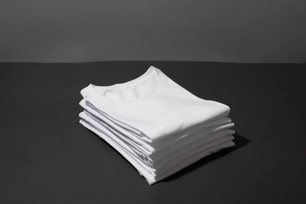 Closeup of a stack of folded white cotton t shirts isolated over dark gray background - Foto, imagen