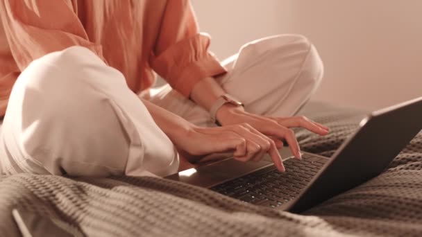 Cropped unrecognizable person wearing white trousers and peachy blouse sitting in lotus position on top of bed, typing on portable computer - Footage, Video