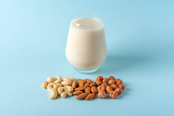 Glass of organic vegan dairy free milk from nuts. Healthy vegetarian alternative drink. Various types of nuts: cashew, hazelnuts, almonds on blue background. Copy space for text - Foto, immagini