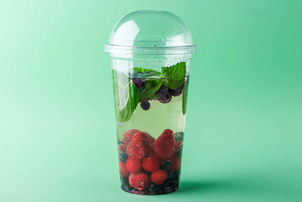 Fresh cool detox drink with Various berries in plastic cup on green background. Tasty infused water or lemonade to go. Proper nutrition and healthy eating. Fitness diet. Copy space for text, for menu - Valokuva, kuva