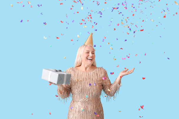 Mujer madura feliz en sombrero de fiesta y con regalo en el fondo de color - Foto, Imagen