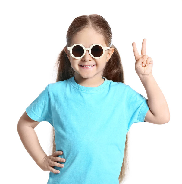 Menina bonito vestindo óculos de sol elegantes e mostrando gesto de vitória no fundo branco - Foto, Imagem