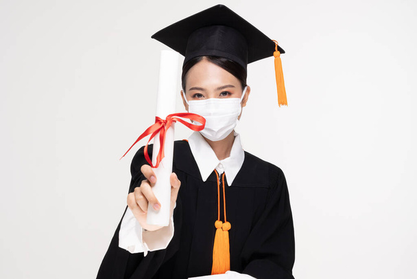 Attractive Asian woman graduated in cap and gown wear medical mask smile so proud and happiness due Virus outbrake and COVID-19 virus flu pandemic on white background,New normal of graduation concept - Foto, Imagen