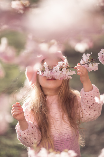 concentrazione selettiva. Una bella ragazza con lunghi capelli biondi in maglione rosa e basco si sta godendo la fioritura delle mandorle. Primavera. Bambino tiene ramoscello, annusa fiori, sorrisi - Foto, immagini