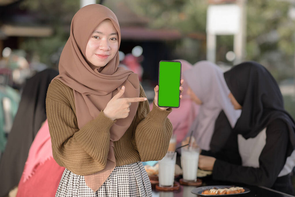 młody Azji nastolatek dziewczyna pokazując i wskazując puste zielony telefon ekran podczas przyjaciele siedzi w tle na kawiarni lub jadłodajni - Zdjęcie, obraz