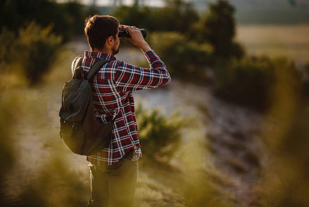 A fickó távcsövet néz a hegyekben. Egy férfi pólóban, hátizsákkal. Fiatal kaukázusi férfi túrázás közben a völgyben táj - Fotó, kép