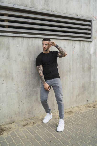 A vertical shot of a stylish European man with tattooed arms leaning on a wall - Фото, зображення