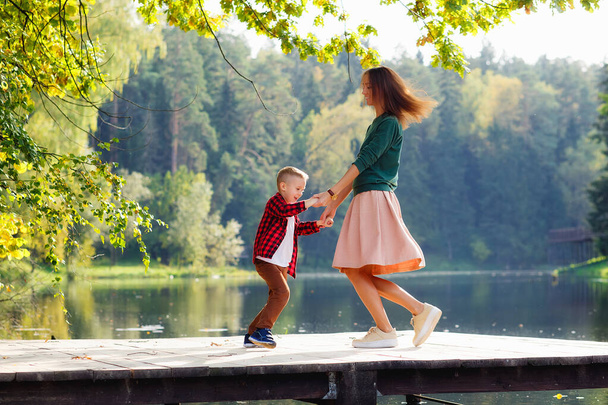 Szczęśliwa matka i syn na drewnianym pomoście nad jeziorem jesienią. Kobieta i dziecko tańczą, wirują, trzymają się za ręce. - Zdjęcie, obraz