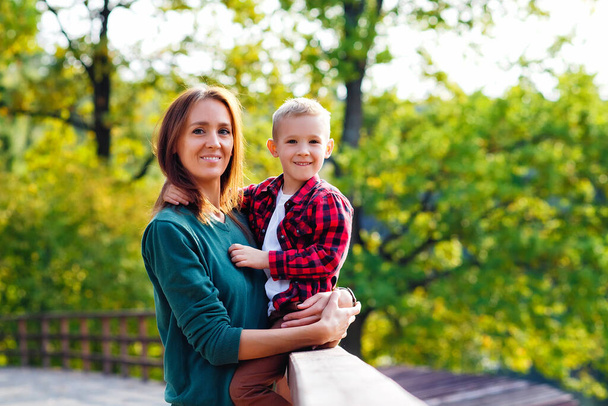 Mooie vrouw en jongen op een houten brug. De zoon in een rood geruit shirt knuffelt zijn moeder zittend op de reling. - Foto, afbeelding