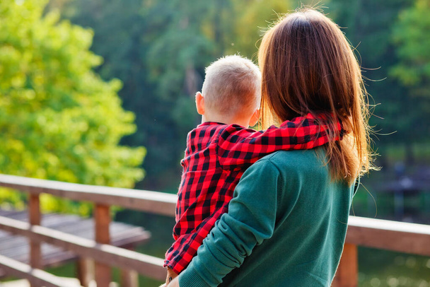 Μια όμορφη γυναίκα και ένα αγόρι σε μια ξύλινη γέφυρα. Η μητέρα κρατά τον γιο της στην αγκαλιά της και στέκεται με την πλάτη της στην κάμερα.. - Φωτογραφία, εικόνα