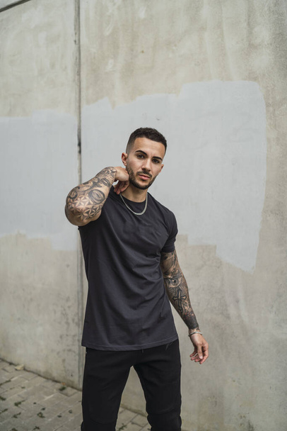 A vertical shot of a stylish European man with tattooed arms posing against a concrete wall - Fotografie, Obrázek