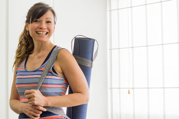 A cheerful Latina woman carrying a yoga mat with a strap on her shoulder - Photo, Image