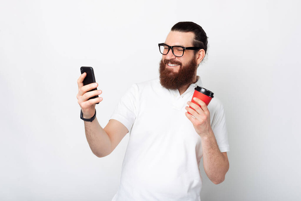 Veselý pohledný vousatý muž pomocí smartphonu a drží šálek kávy přes bílé pozadí. - Fotografie, Obrázek