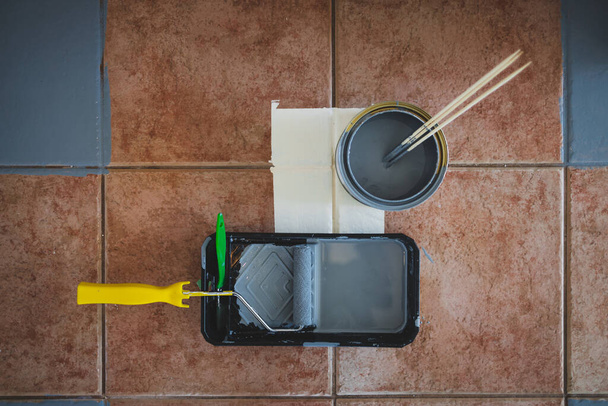paint roller with brush and tray next to open pot of paint shot from above surrounded by terracotta floor tiles being painted gray, diy and home renovation concept - Фото, зображення