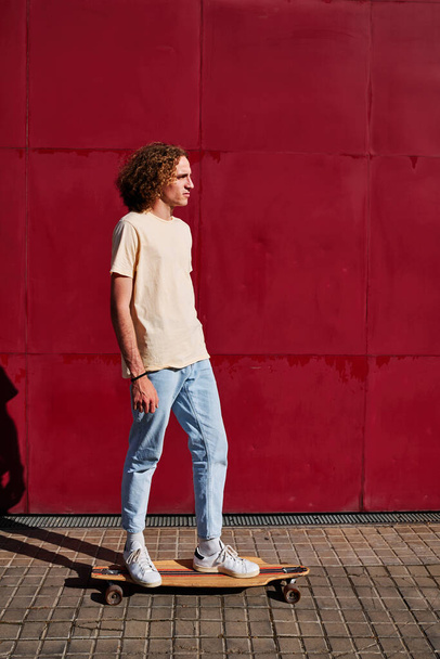 a young man with curly hair using his skateboard with a red background behind him on a sunny day - Fotó, kép