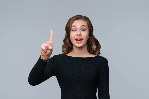 Młoda, pozytywna brunetka wskazująca palcem w górę, pokazująca reklamę na górze i uśmiechnięta, promująca firmowy sztandar, stojąca na szarym tle. Ludzkie emocje, koncepcja gestu - Zdjęcie, obraz