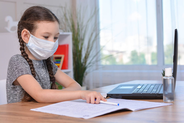 Een schoolmeisje met een medisch masker doet haar huiswerk op een laptop thuis of studeert in de klas tijdens een les op school.Studentenveiligheid tijdens de coronavirusuitbraak. Terug naar school.Afstandsonderwijs. - Foto, afbeelding