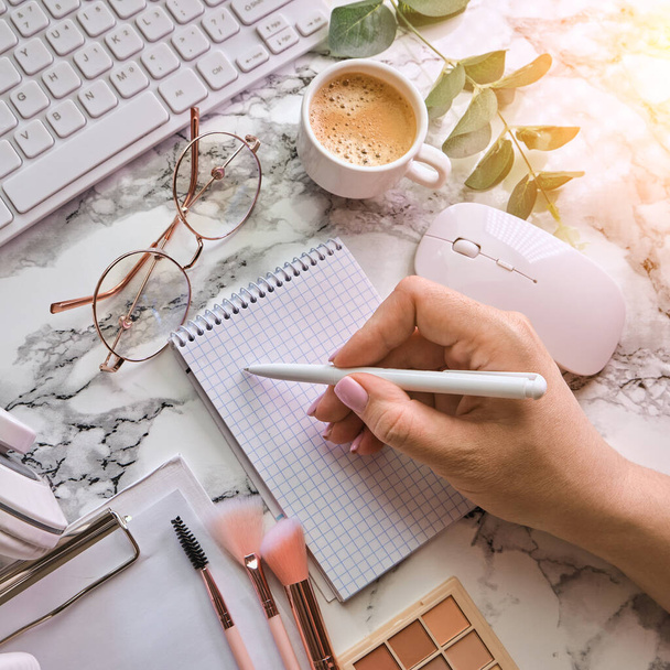 Workplace mockup with female hand writing, notebook, glasses, headphones and make up accessories on white marble background top view. Feminine working style concept. - Photo, Image