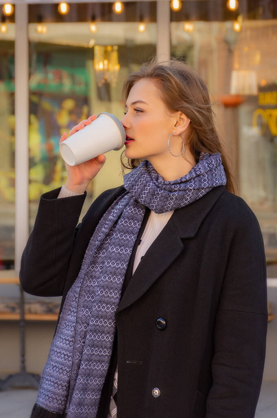 Młoda uśmiechnięta kobieta w czarnym płaszczu i szaliku trzymająca kawę z ekologicznym kubkiem wielokrotnego użytku na miejskim tle gabloty. Koncepcja przyjazna środowisku. - Zdjęcie, obraz
