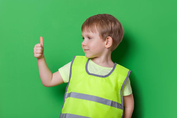 Καυκάσιος παιδί αγόρι σε ανοιχτό χρώμα φωτεινό γιλέκο με ανακλαστικές λωρίδες δείχνει αντίχειρες προς τα πάνω χειρονομία, μεγάλη, και χαμόγελο. Στούντιο γυρίστηκε σε πράσινο φόντο, την ασφάλεια και την κυκλοφορία των κανόνων έννοια. - Φωτογραφία, εικόνα