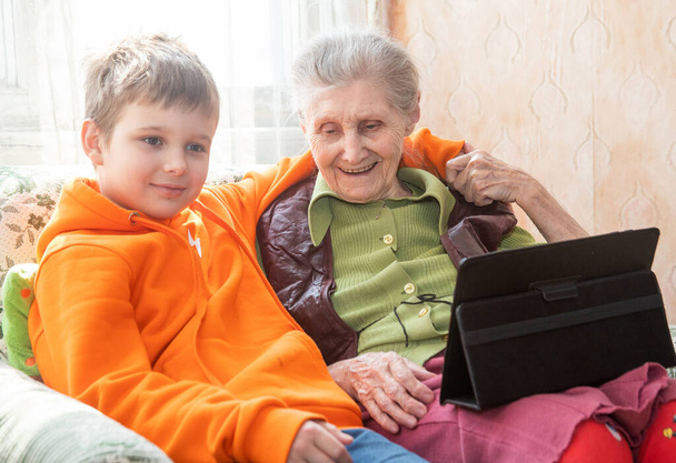 Une vieille femme européenne avec un garçon mignon dans un raglan orange est assis avec une tablette et bavarder en ligne. - Photo, image